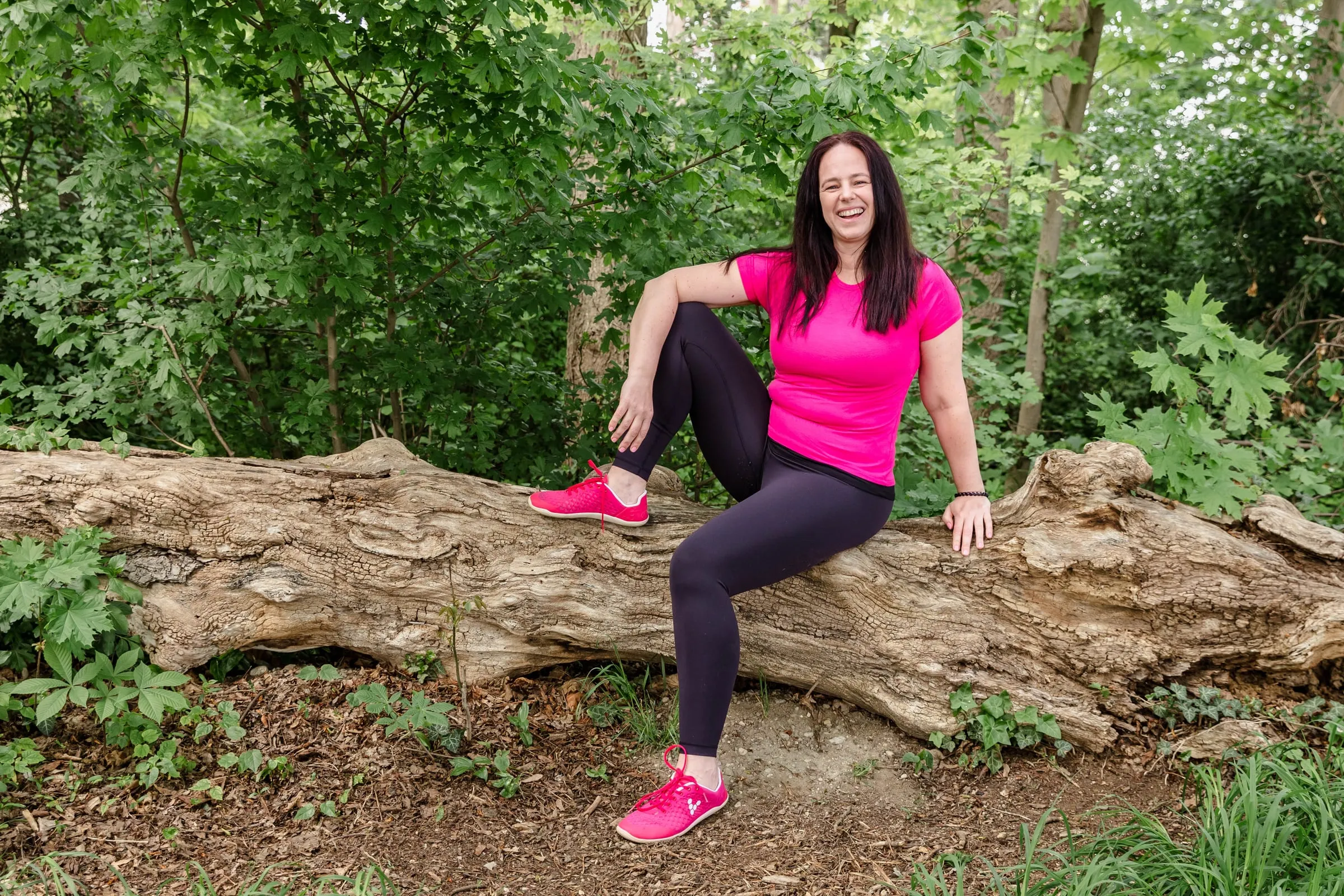 Madlen in Pink sitzt lachend auf einem Baumstamm im Wald
